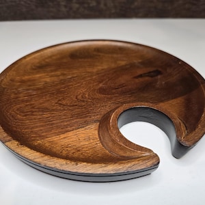 Może przedstawiać: Drewniana taca do serwowania z ciemnobrązowym wykończeniem i wycięciem w kształcie półksiężyca. Taca ma gładką, wypolerowaną powierzchnię i czarną obwódkę. Ta ręcznie robiona taca jest idealna do serwowania przekąsek lub napojów.
