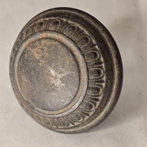 May include: A round, dark metallic cabinet knob with a weathered, aged finish. The knob features a smooth, flat circular center surrounded by a raised, textured border with a repeating arched pattern along the outer edge of the hardware piece.