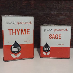 Może przedstawiać: Dwa vintage metalowe pojemniki na przyprawy z czerwonymi i białymi etykietami. Większy pojemnik jest oznaczony jako "pure ground THYME", a mniejszy jako "pure ground SAGE". Oba pojemniki mają czarno-białe logo w kształcie łzy z napisem "Tone's" wewnątrz.