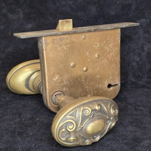 Op de afbeelding: Een messing insteekslot met een rechthoekig mechanisme, een bovenste dagschoot en twee ovale deurknoppen. De knoppen zijn voorzien van een reliëf met krulmotieven en het geheel vertoont een verweerde patina.