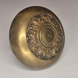 Może przedstawiać: Zbliżenie na zabytkową mosiężną gałkę do drzwi. Gałka ma zaokrąglony kształt z ozdobnym, okrągłym wzorem pośrodku. Wzór zawiera szczegółowe wzory kwiatowe i zwoje. Mosiądz ma ciepłą, postarzaną patynę.