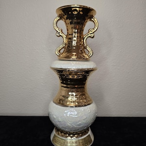 Puede incluir: Un jarrón decorativo de cerámica con la parte superior y detalles dorados, y una base blanca con motivos florales en relieve. El jarrón tiene una forma única con dos asas en la parte superior. La base tiene el texto "BEAM'S KENTUCKY STRAIGHT BOURBON WHISKEY".