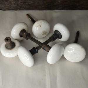 May include: A collection of five white porcelain door knobs with dark metal spindles and bases. The knobs are round and smooth, showing signs of age and wear on the surfaces. Each knob is attached to a square metal shaft, with some pieces connected in pairs and others appearing as individual units.