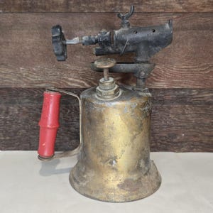 Op de afbeelding: Een vintage messing soldeerbrander met een cilindrische brandstoftank, een zwarte metalen branderkop met een regelventiel en een rode cilindrische handgreep. Het messing oppervlak vertoont tekenen van ouderdom en oxidatie.