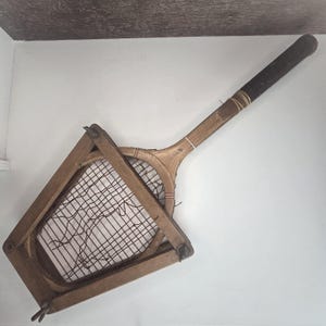 May include: An antique wooden tennis racket with a dark grip and a rectangular frame. The racket features a classic design with a wooden handle and a stringed net. The frame is made of wood and is connected to a rectangular wooden frame.