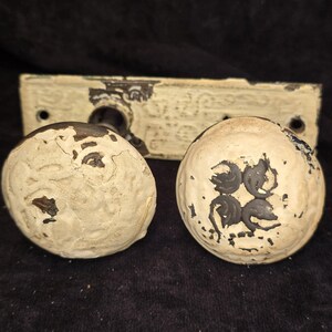 May include: A pair of round, off-white door knobs with a distressed, chipped paint finish revealing dark metal underneath. The knobs feature a raised, embossed floral pattern and are attached to a rectangular, matching off-white backplate.