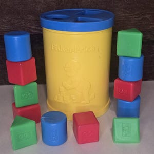 Pode incluir: Um brinquedo vintage de classificação de formas Fisher-Price. O recipiente amarelo tem uma tampa azul e uma figura de criança em relevo na lateral. Blocos vermelhos, azuis e verdes sortidos em várias formas estão espalhados ao redor do recipiente. O texto "Fisher-Price" é visível.