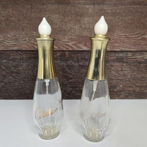 Könnte beinhalten: Zwei Vintage-Parfümflaschen mit transparenten Glaskörpern und goldfarbenen Metallakzenten. Jede Flasche hat einen weißen, tropfenförmigen Verschluss und den Schriftzug "Corallion" in Gold. Der Hintergrund ist aus Holz.