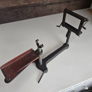 May include: An antique, black metal device with a rectangular frame and a red-brown tray. The device has a long arm and a vertical support with a clamp. The frame is rectangular and appears to be adjustable. The device is likely used for a specific purpose.