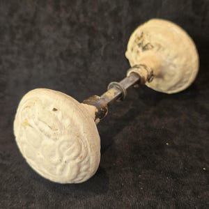 May include: A pair of round, off-white door knobs with a textured, embossed floral pattern on the surface. The knobs are connected by a central metal spindle with a dark, weathered finish, showing signs of age and wear on the hardware components.