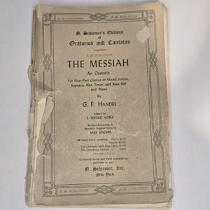 Pode incluir: Uma partitura musical vintage intitulada "THE MESSIAH" de G.F. Handel. A capa, com seu fundo creme e texto preto, mostra sinais de desgaste. Os detalhes da editora, "G. Schirmer, Inc., New York", também são visíveis.