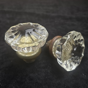 Puede incluir: Dos pomos de puerta de cristal transparente con un diseño facetado. Los pomos tienen forma redonda y están montados sobre una base de color latón. El cristal es transparente, permitiendo el paso de la luz. Los pomos son probablemente vintage o antiguos.