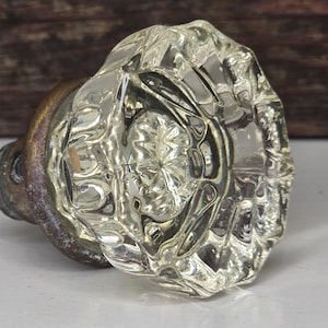 May include: A clear, faceted glass doorknob with a brass base. The knob has a rounded shape with a detailed, starburst-like pattern in the center. The brass base shows signs of age and wear.
