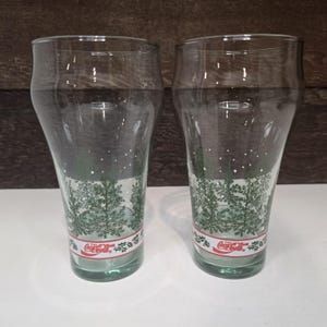 May include: Two clear Coca-Cola pint glasses with a winter forest design. The glasses feature a band of green trees and falling snow, with the Coca-Cola logo in red on a white band at the base. The glasses are approximately 6 inches tall.