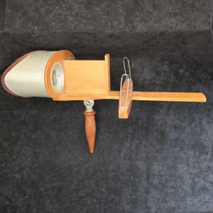 May include: A vintage stereoscope viewer featuring a light brown wooden body with a textured finish, a silver-toned metal hood with two circular viewing lenses, a dark brown wooden handle, and a sliding wooden card holder with metal wire clips.