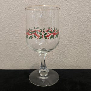 May include: A clear glass wine goblet with a gold rim and a decorative band of red ribbons, green holly leaves, and red berries. The stem is clear and the base is round.