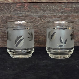 May include: Two frosted glass tumblers with a silver band and a frosted leaf design. The glasses are on a black surface.