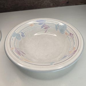 May include: A round, off-white ceramic bowl with a floral design. The bowl features a light blue rim and a decorative pattern of blue, purple, and peach-colored flowers. The bowl has a slightly textured surface.