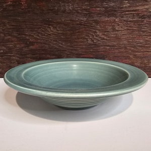 Könnte beinhalten: Eine türkisfarbene Keramikschale mit breitem, flachem Rand und leicht konkavem Zentrum. Die Schale hat horizontale Rillen um den Boden. Der Hintergrund ist eine dunkle Holzoberfläche.