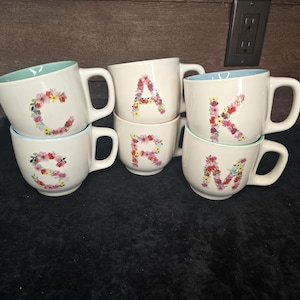 Pode incluir: Seis canecas de cerâmica branca com desenhos florais que soletram a palavra "CARM". Cada caneca tem uma borda de cor diferente, incluindo azul, rosa e verde.