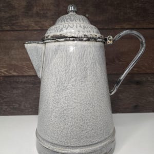 May include: A vintage gray and white enamelware coffee pot with a lid and handle. The pot has a speckled pattern and a spout for pouring. The handle is a curved metal. The pot is sitting on a white surface.