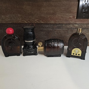May include: Four vintage glass aftershave bottles with different designs. The bottles are brown and black with gold accents. The bottles are arranged in a row on a wooden surface.