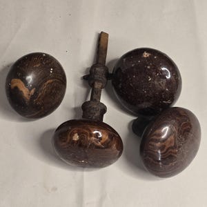 Op de afbeelding: Vier ronde deurknoppen met een donkerbruin en beige marmerpatroon. Drie knoppen zijn los, terwijl één knop vastzit aan een vierkante metalen spindel met een schroefkraag en een kleine metalen bevestiging aan de basis van de knop.