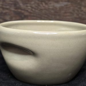 May include: A light beige ceramic bowl with a unique, asymmetrical shape. The bowl has a smooth, slightly textured surface and a subtle, uneven glaze.