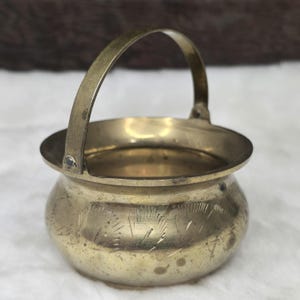 Könnte beinhalten: Eine kleine, dekorative Schale aus Messing mit einem Henkel. Die Schale hat eine runde, leicht ausgestellte Form und ist mit einem sich wiederholenden Muster aus geätzten Linien verziert.