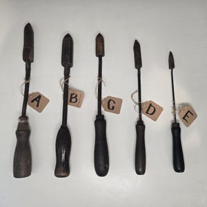 Puede incluir: Se muestran cinco cautines antiguos, cada uno con mango de madera y punta de metal. Los cautines varían en forma y tamaño, con etiquetas de la A a la E. Están dispuestos verticalmente sobre un fondo blanco, resaltando su aspecto envejecido.