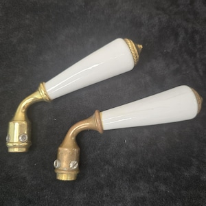 Puede incluir: Dos manijas de puerta antiguas con palancas de porcelana blanca y herrajes de latón. Las manijas tienen un diseño curvo y están montadas sobre un fondo oscuro. Las palancas son cónicas y tienen un acento decorativo de latón en el extremo.