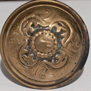 Puede incluir: Pomo de puerta antiguo de latón con un diseño floral en relieve. El pomo circular presenta un medallón central rodeado de volutas detalladas y un borde de cuentas. El acabado envejecido le da una estética vintage.