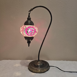 May include: A pink and white mosaic glass lamp with a curved metal arm and a brass base. The lamp is turned on and the light shines through the mosaic glass.