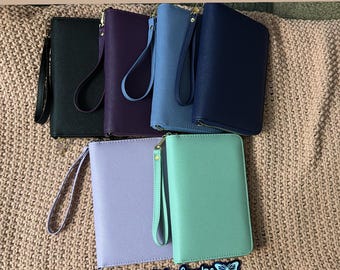 May include: Six different colored zippered wallets with wrist straps. The wallets are arranged in a row on a beige knitted surface. The wallets are black, purple, light blue, dark blue, lavender, and light green.