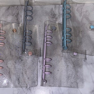 May include: Five clear plastic binders with glitter embedded in the plastic. The binders have different coloured rings: gold, grey, purple, blue, and pink.