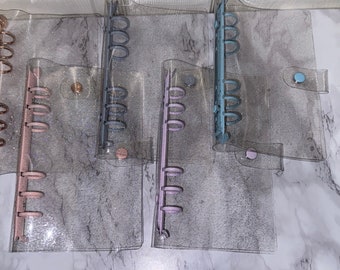 May include: Five clear plastic binders with glitter embedded in the plastic. The binders have different colored rings: gold, gray, purple, blue, and pink.