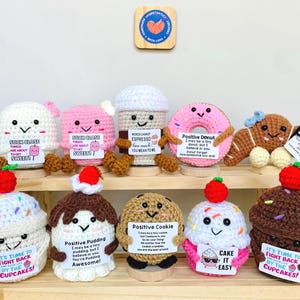 May include: A collection of crocheted food items, including a white ghost, pink ghost, coffee cup, pink donut, gingerbread person, cupcake, pudding, cookie, and a chocolate cupcake. Each item has a smiling face and a small sign with a positive message.