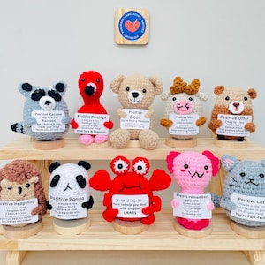 May include: A set of 10 crocheted animal plushies, each with a positive message. The plushies are arranged on a two-tiered wooden shelf. The animals include a hedgehog, raccoon, flamingo, bear, cow, otter, panda, crab, axolotl, and cat.