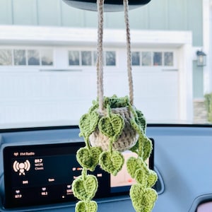 Puede incluir: Decoraci&oacute;n de planta colgante de ganchillo para coche. La maceta es beige, con hojas verdes en cascada. La planta cuelga de dos cordones beige sujetos al espejo retrovisor.