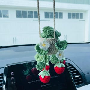 Puede incluir: Un adorno de coche de fresa hecho a ganchillo. El adorno presenta una cesta beige con hojas verdes, flores blancas y fresas rojas. Cuelga de una cuerda beige y est&aacute; suspendido frente al salpicadero de un coche.