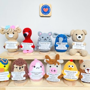 May include: A collection of handmade crochet animal figurines, including a cow, flamingo, cat, elephant, bear, duck, hedgehog, shark, cow, chick, and otter. Each holds a sign with an inspirational message. The figurines are displayed on wooden stands.