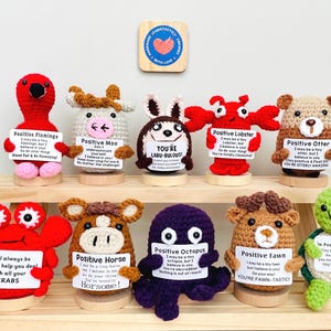 May include: A collection of ten crocheted animal figurines, each holding a sign with an inspirational message. The animals include a flamingo, cow, bunny, lobster, otter, crab, horse, octopus, fawn, and turtle. The figurines are in various colors and sit on a wooden shelf.