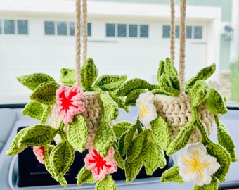 May include: Two crocheted flower arrangements hanging from the rearview mirror of a car. The arrangements are made with green leaves, white and pink flowers, and a beige crocheted pot.