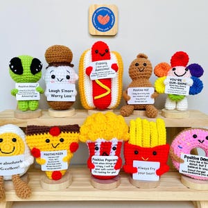 May include: A collection of handmade crochet food items, including an alien, s'more, hot dog, nut, flower, egg, pizza, popcorn, french fries, and donut. Each item holds a sign with a positive message. The items are displayed on a wooden shelf.