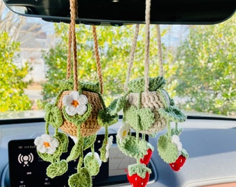 May include: Three crocheted hanging ornaments in a car. The ornaments are shaped like pots with flowers and leaves hanging from them. The pots are brown, beige, and green. The flowers are white and the leaves are green. The ornaments are hanging from the rearview mirror of a car.