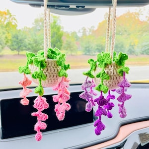 Puede incluir: Dos decoraciones colgantes de plantas de ganchillo. Cada una presenta una maceta beige con hojas verdes y flores rosas o moradas en cascada. Las plantas est&aacute;n suspendidas por cuerdas de color crema, a&ntilde;adiendo un toque de encanto hecho a mano.