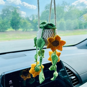 Puede incluir: Amuleto de coche de ganchillo hecho a mano con un dise&ntilde;o de maceta colgante, con girasoles amarillos y hojas verdes. La maceta es blanca y beige, suspendida del espejo retrovisor.