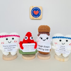 May include: Four crocheted plush toys, each with a different design and a message. The first is a white tooth with pink trim holding a sign that says "You're awesome, and that's the tooth!" The second is a red watermelon with a sign that says "Positive Watermelon will always be around to let you know that you are one in a MELON." The third is a brown crocheted person with a sign that says "Laugh S'more Worry Less." The fourth is a white tooth with blue trim holding a sign that says "You're Toothy Amazing! Be the reason someone smiles today."