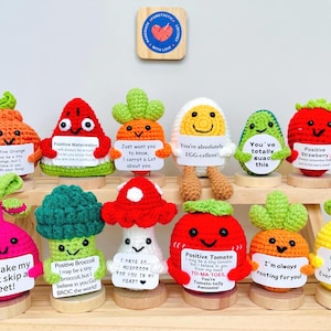 May include: A collection of handmade crochet food plushies, including an orange, watermelon, carrot, egg, avocado, strawberry, beet, broccoli, mushroom, tomato, and apple. Each plushie holds a sign with a positive message. A wooden sign with a heart is at the top.