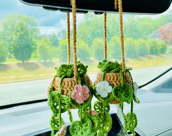 May include: Two crocheted hanging plants with green leaves and pink and white flowers, hanging from the rearview mirror of a car.